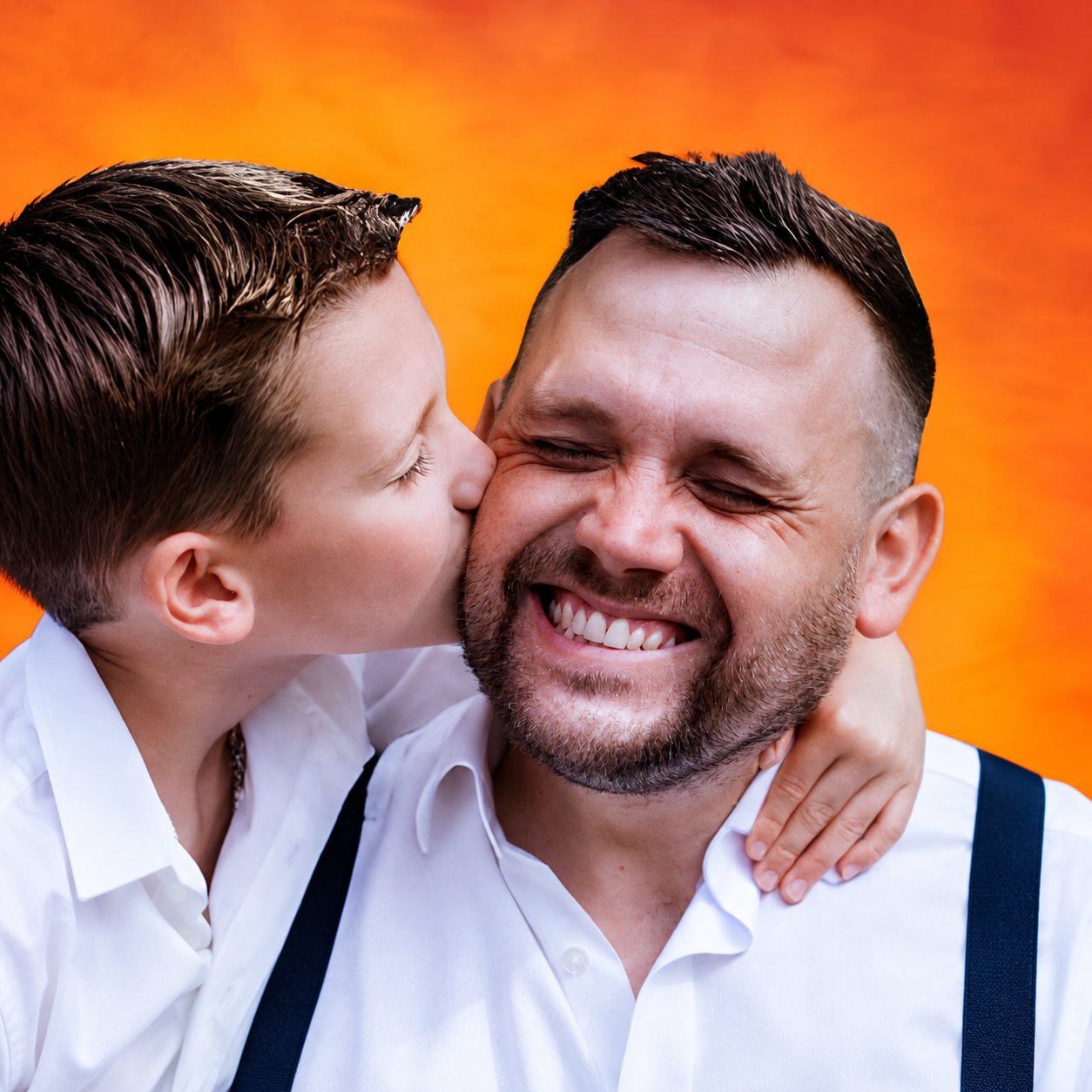 AJ Ferguson in a white shirt and navy suspenders, smiling as his young son kisses him on the cheek against a vivid orange backdrop